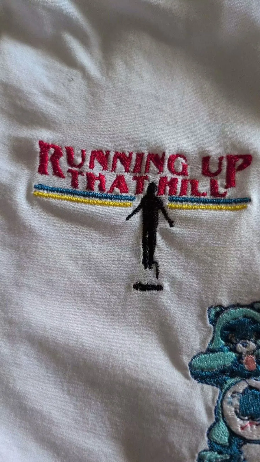 White fabric with embroidered ‘Running Up That Hill’ lettering, coloured lines and a small figure