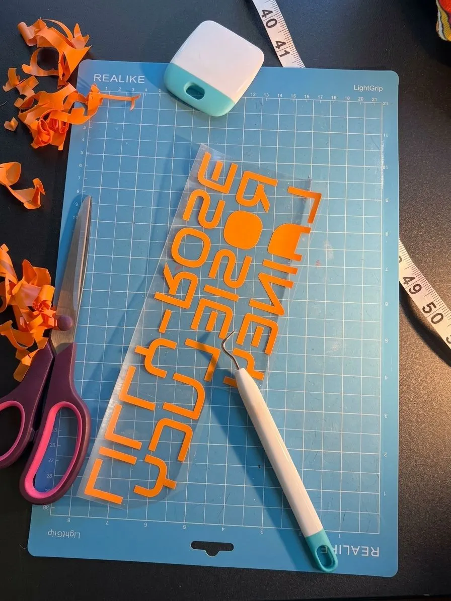 Overhead studio photograph of orange vinyl lettering on transfer tape on a blue Realike cutting mat with scissors, a weeding tool and offcuts