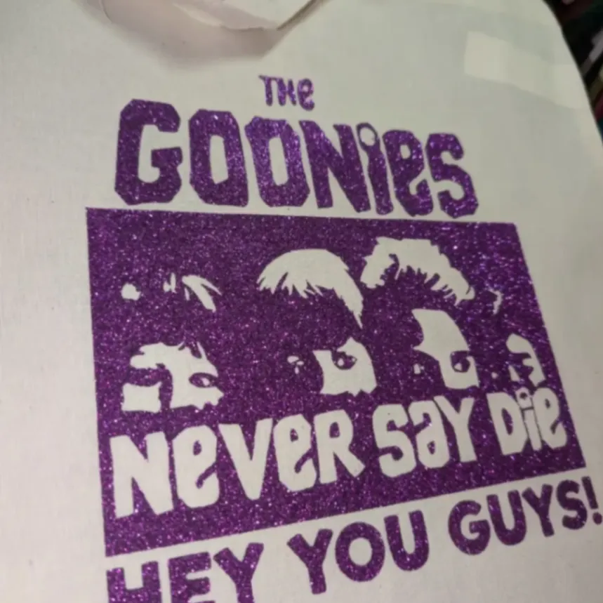 Cream tote bag with a purple The Goonies Never Say Die design and character silhouettes